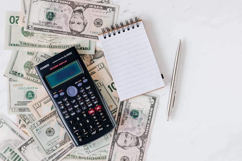 Calculator, blank notepad, and pen resting on a pile of U.S. dollar bills on a white surface.